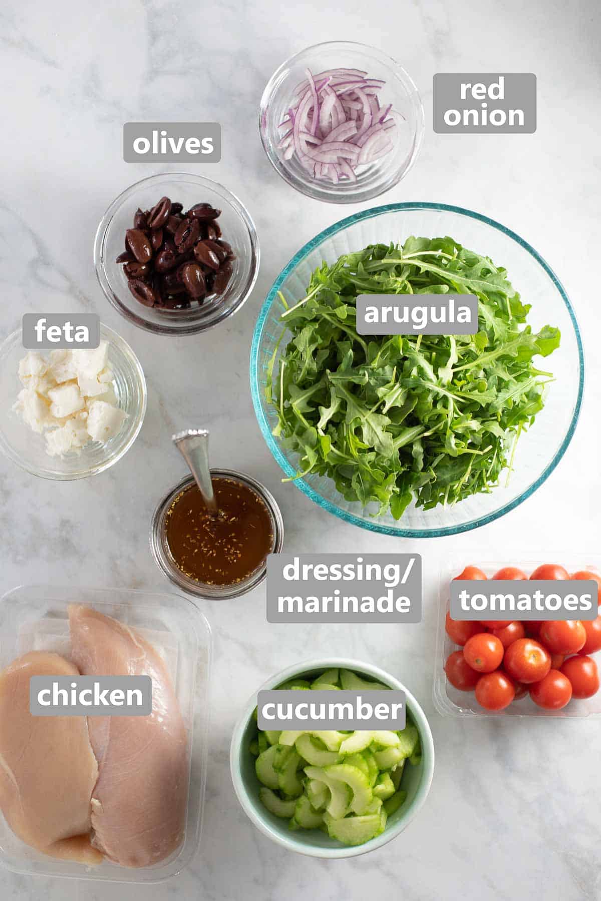 Mediterranean salad ingredients in glass bowls along with raw chicken breasts