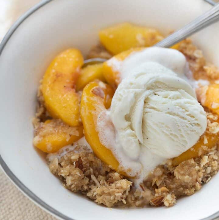 close up shot of peach crisp in white bowl topped with ice cream and spoon