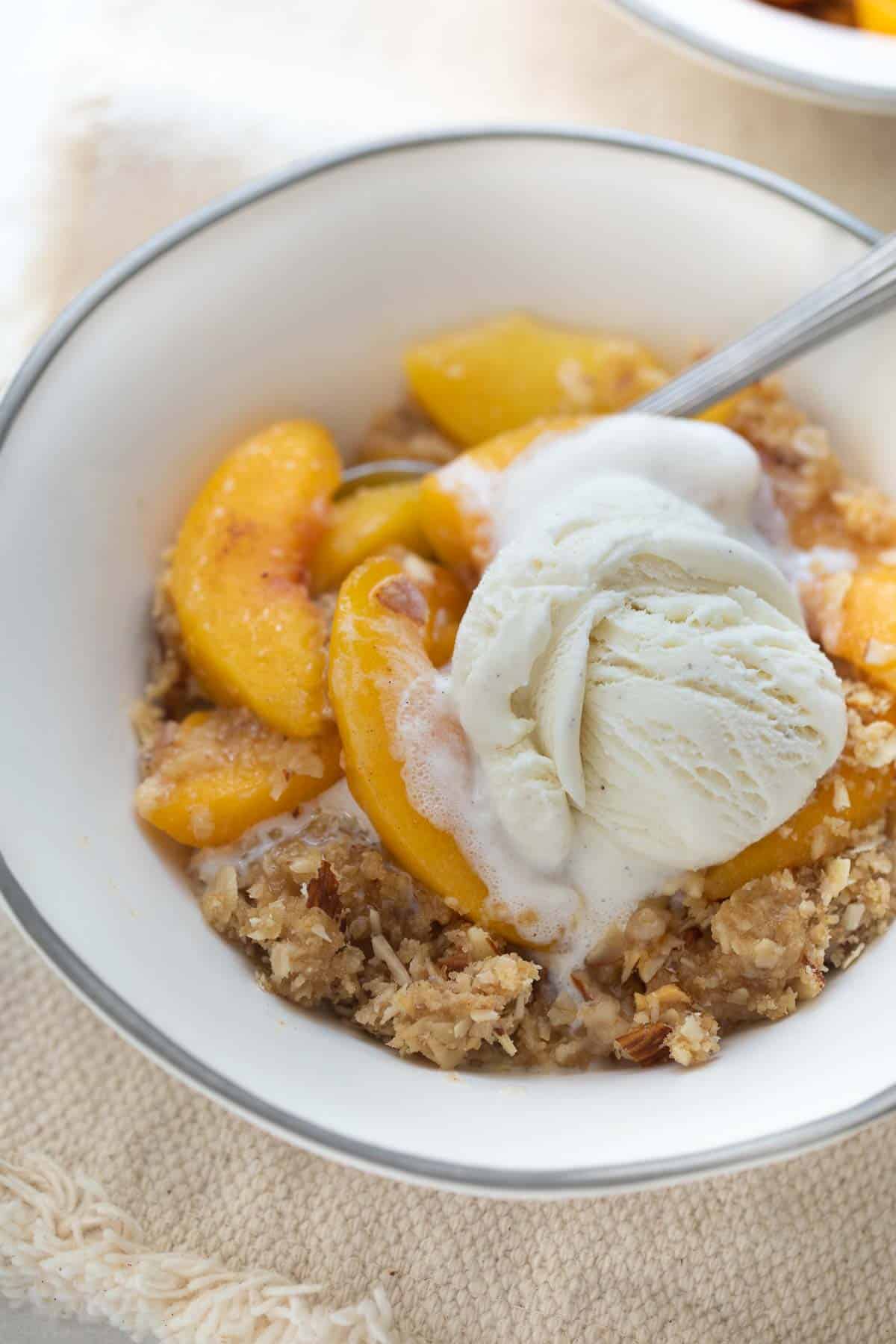 close up shot of peach crisp in white bowl topped with ice cream and spoon