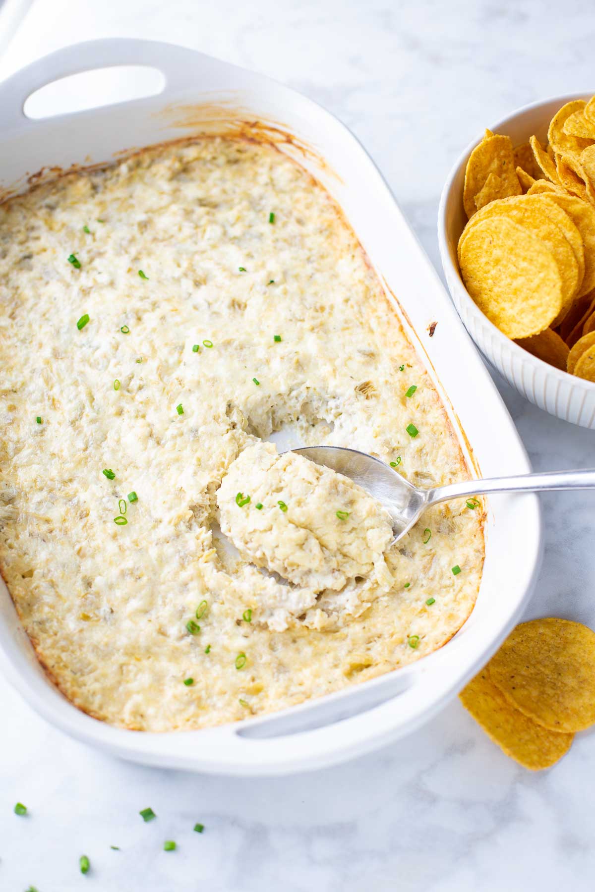 white casserole dish with artichoke dip and serving spoon next to bowl of tortilla chips