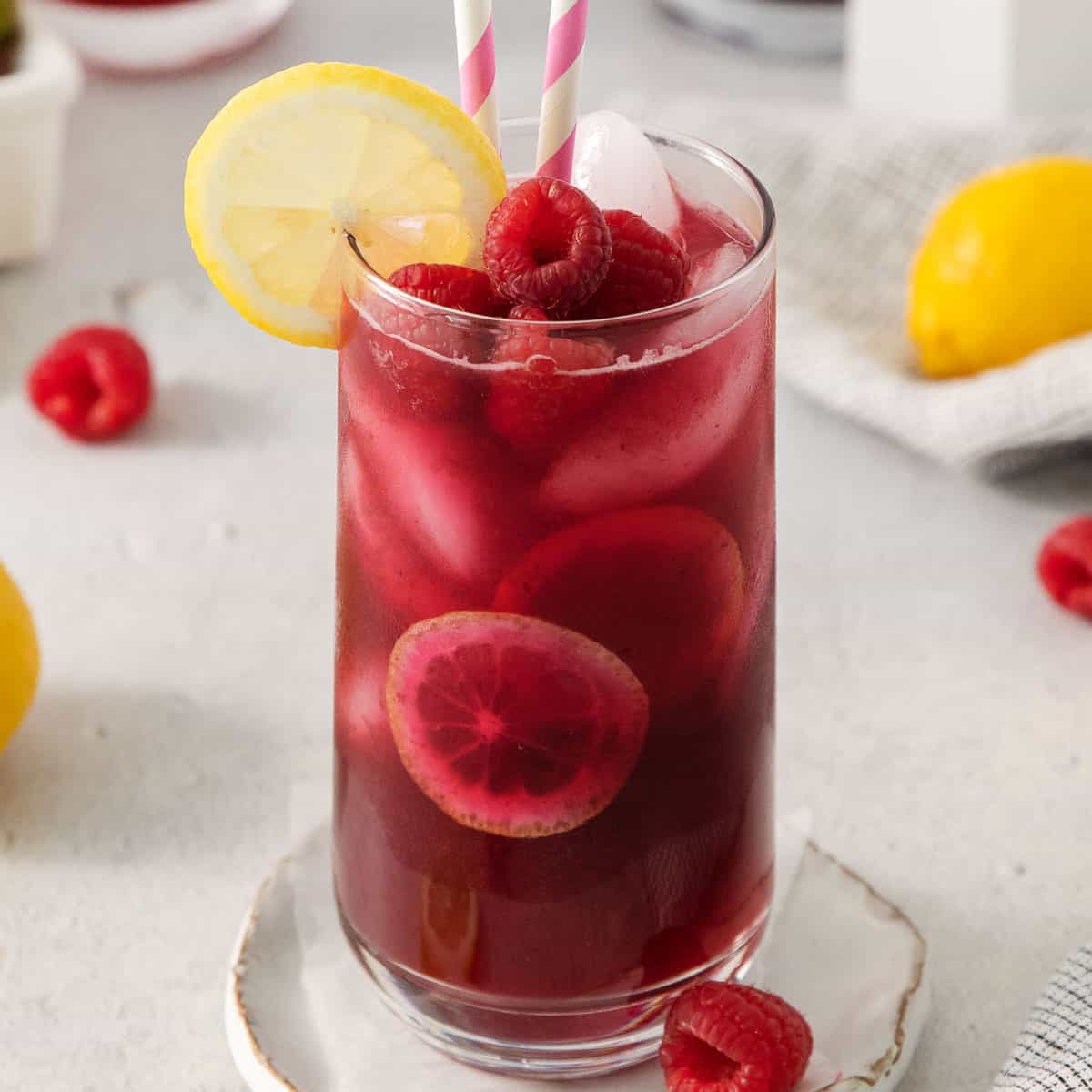 A close up photo of berry lemonade in a glass with two striped straws and a slice of lemon on the side