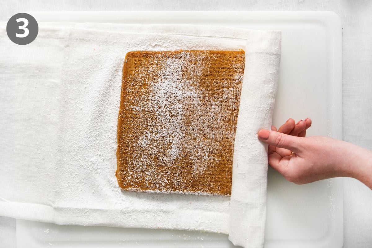 A hand uses a kitchen towel to roll a rectangular, powdered sugar-dusted cake sponge on a cutting board.