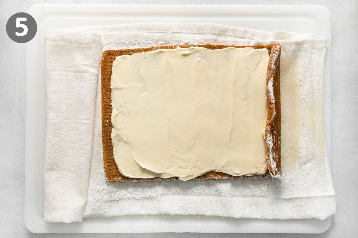 A rectangular sponge cake layer covered in white frosting, resting on a dusted kitchen towel on a cutting board.