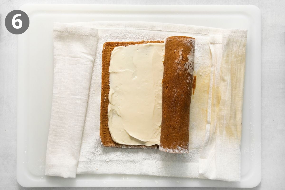 A rectangular cake sponge spread with cream being rolled up on a towel dusted with powdered sugar.
