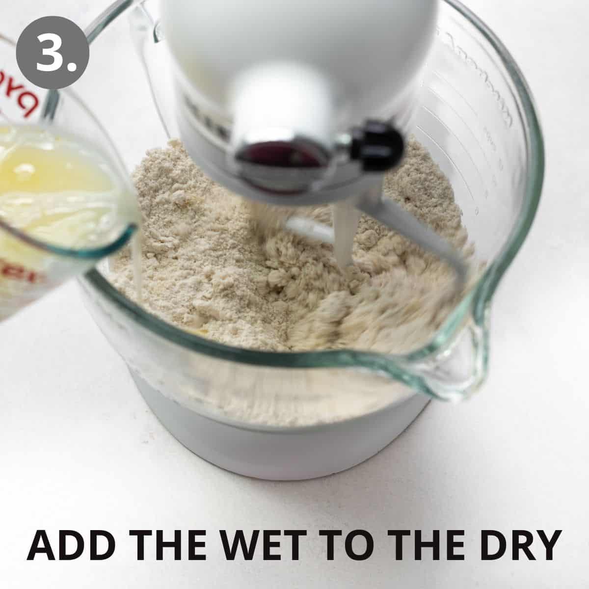 A standing mixer with dry ingredients, and a measuring cup of wet ingredients being poured into the bowl