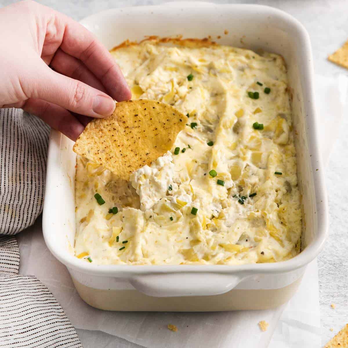 A hand dipping a chip into the dish of hot artichoke dip