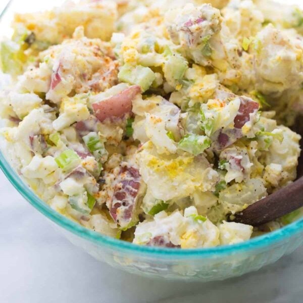 A close-up of a bowl of potato salad with chopped red potatoes, celery, hard-boiled eggs, mayonnaise, and sprinkled seasoning, with a wooden spoon partially visible.