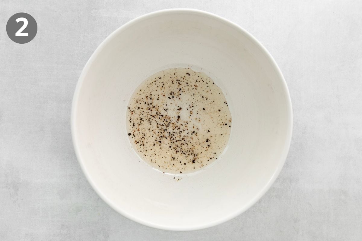 A large white bowl containing a small amount of liquid and ground black pepper, with a gray number 2 in a circle at the top left corner on a light gray background.