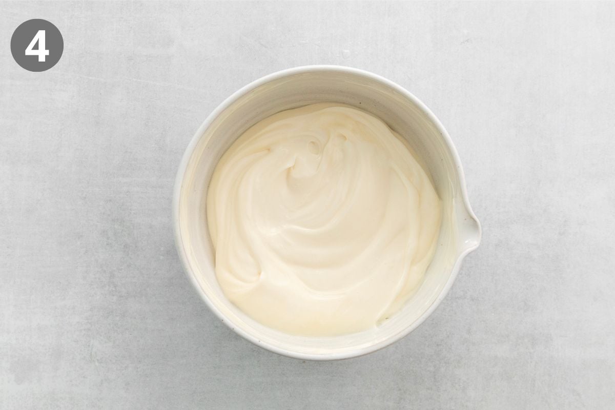 A white mixing bowl filled with smooth, creamy white frosting, shown from above on a light gray surface. A gray circle with the number 4 is in the upper left corner.