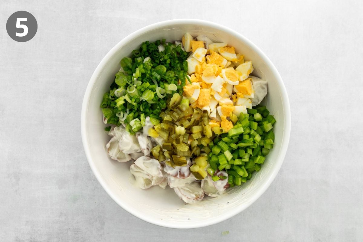 A white bowl containing chopped green onions, hard-boiled eggs, pickles, and celery arranged in separate sections on top of a creamy potato salad. The number 5 is in a circle in the upper left corner.