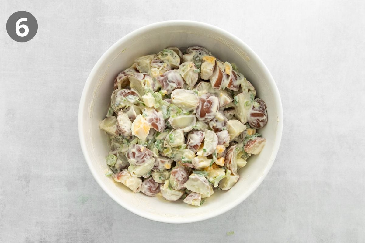 A white bowl filled with creamy potato salad with chunks of potatoes, red grapes, and herbs sits on a light gray surface. A number 6 is displayed in a gray circle in the top left corner of the image.