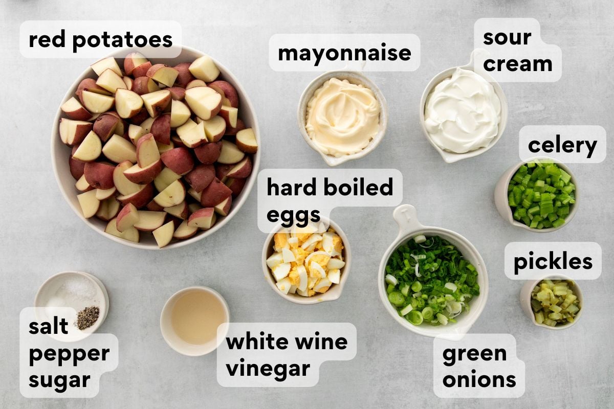 Overhead view of ingredients for potato salad, each labeled: red potatoes (diced), mayonnaise, sour cream, celery (chopped), pickles (chopped), green onions (sliced), hard boiled eggs (chopped), white wine vinegar, salt, pepper, and sugar.