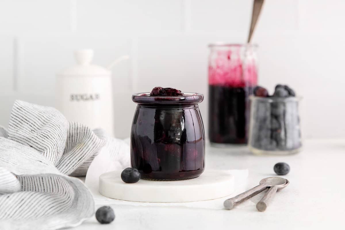 Blueberry compote in a jar, with teaspoons and measuring cups surrounding the jar