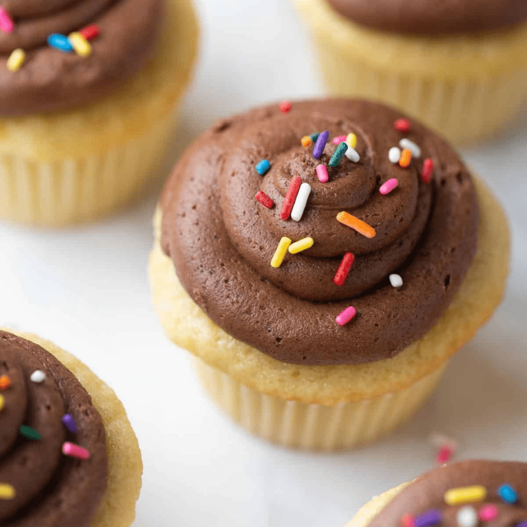Yellow cupcakes lined up on a counter, with chocolate frosting and colorful sprinkles