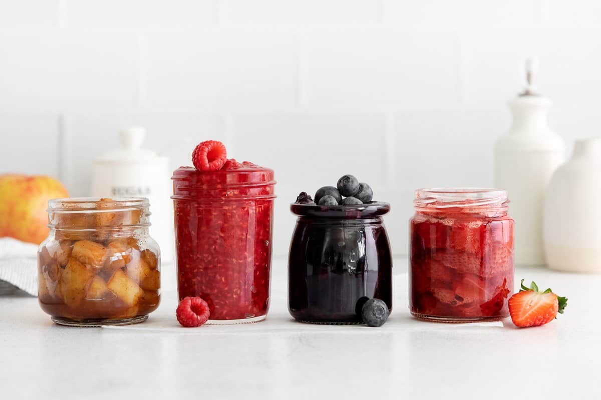 Apple, raspberry, blueberry, and strawberry fruit compote arranged in glass jars on a countertop