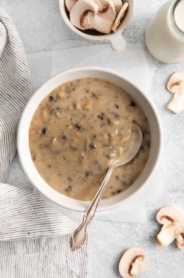 Gluten-free cream of mushroom soup in a bowl with a spoon in it and raw, sliced mushrooms around it