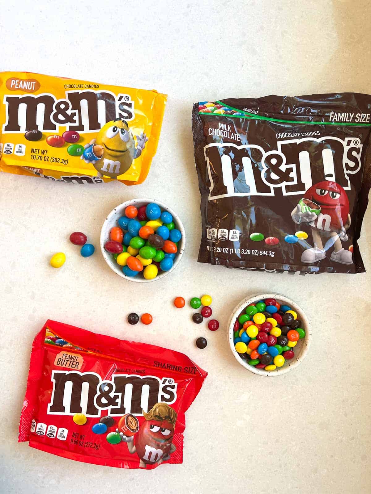 3 bags of M&Ms on white counter with 2 bowls of open mms