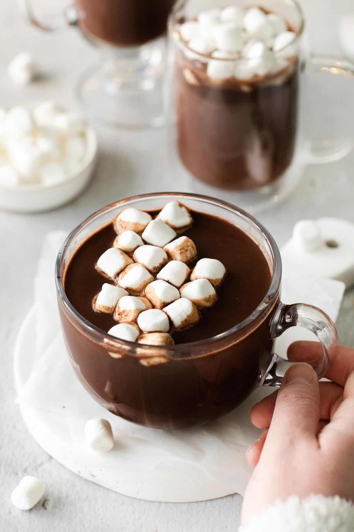 A hand holding a glass mug of hot chocolate with marshmallows on top
