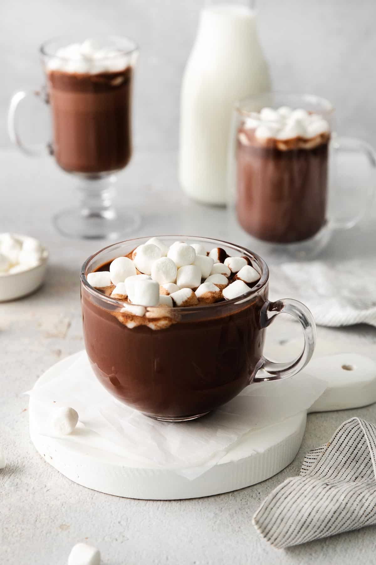 A glass mug of hot chocolate with marshmallows on top, and more mugs of hot chocolate in the background