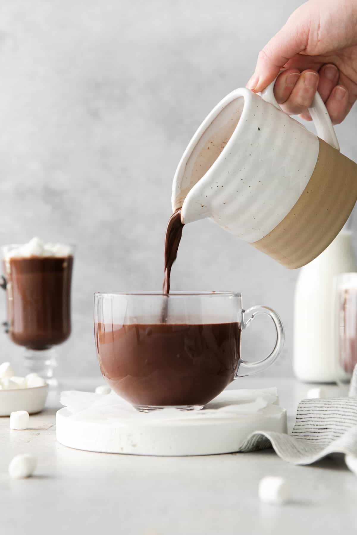 A hand pouring hot chocolate into a glass mug