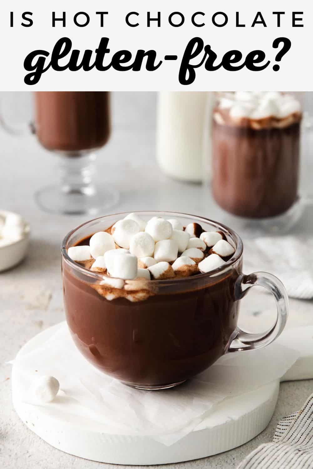 A glass mug of hot chocolate with marshmallows on top, and more mugs of hot chocolate in the background