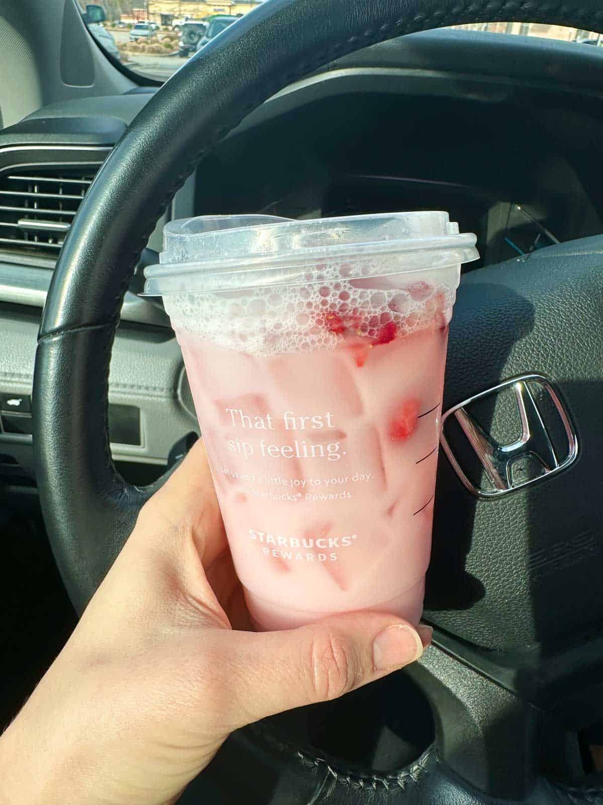 close up shot of starbucks pink drink in hand in front of steering wheel
