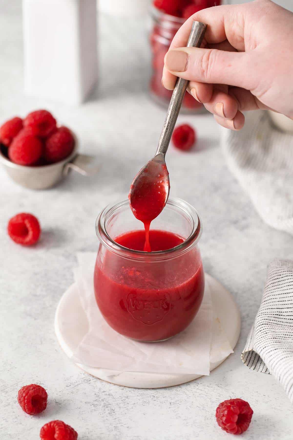 A hand holding a spoon and dipping it into a jar of raspberry coulis