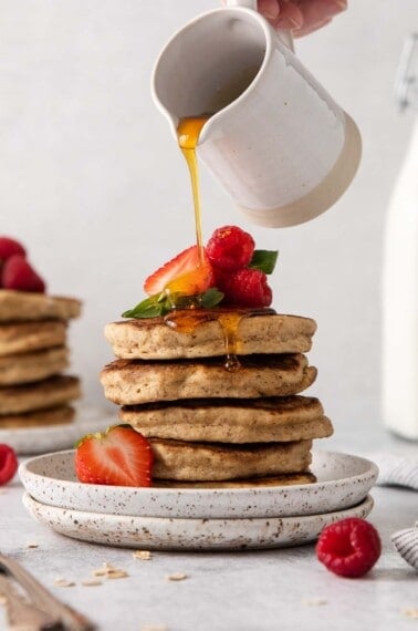 Oat flour pancakes piled on a plate with a hand pouring syrup over the top