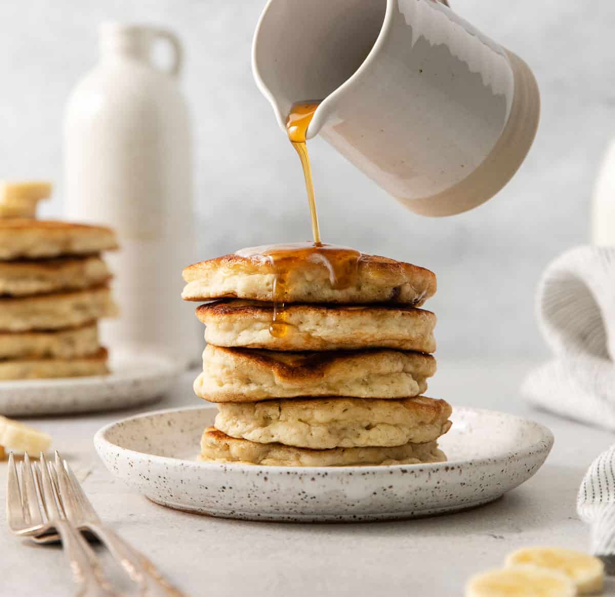Gluten-free banana pancakes stacked up on a plate with a hand pouring syrup over them