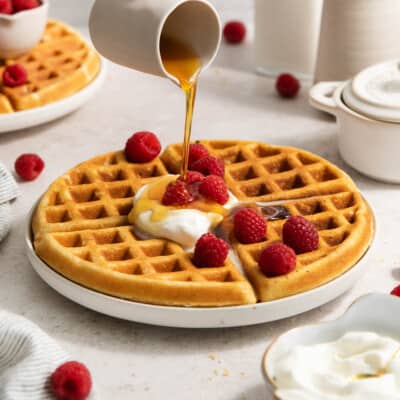 gluten-free waffles on a plate with whipped cream and raspberries and syrup poured over the top