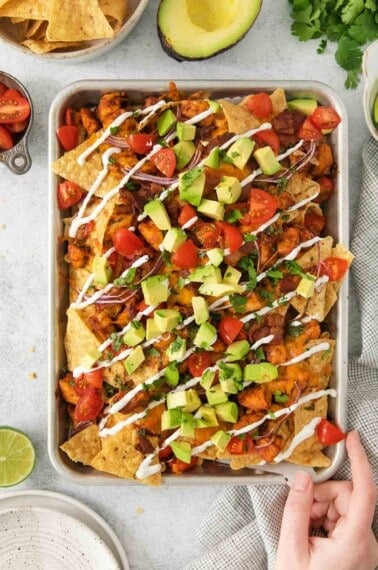BBQ chicken nachos on a sheet pan with a hand touching the pan