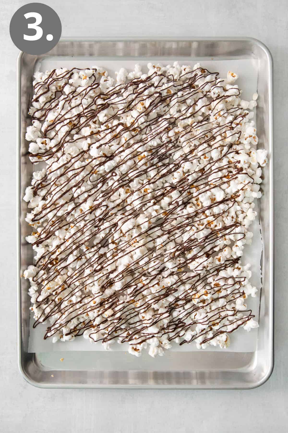Melted chocolate drizzled over popcorn on a baking sheet
