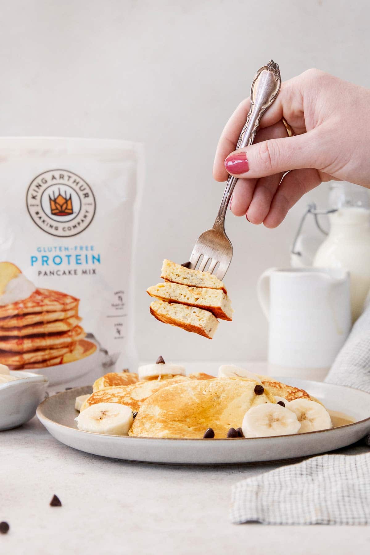 Gluten-free banana protein pancakes on a plate, with a hand taking a bite with a fork and King Arthur Gluten-Free Protein Pancake mix in the background.
