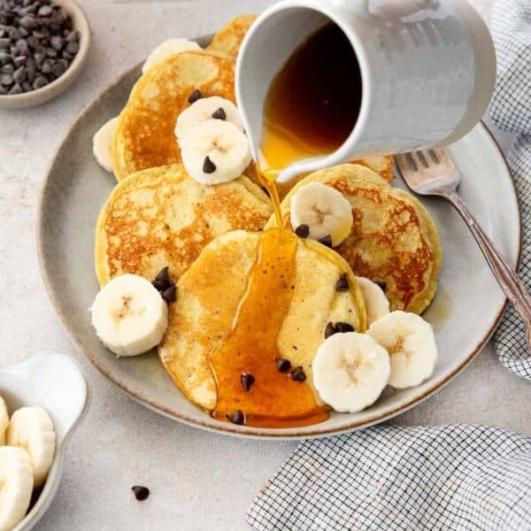 Gluten-free banana protein pancakes on a plate with maple syrup being poured over the top.