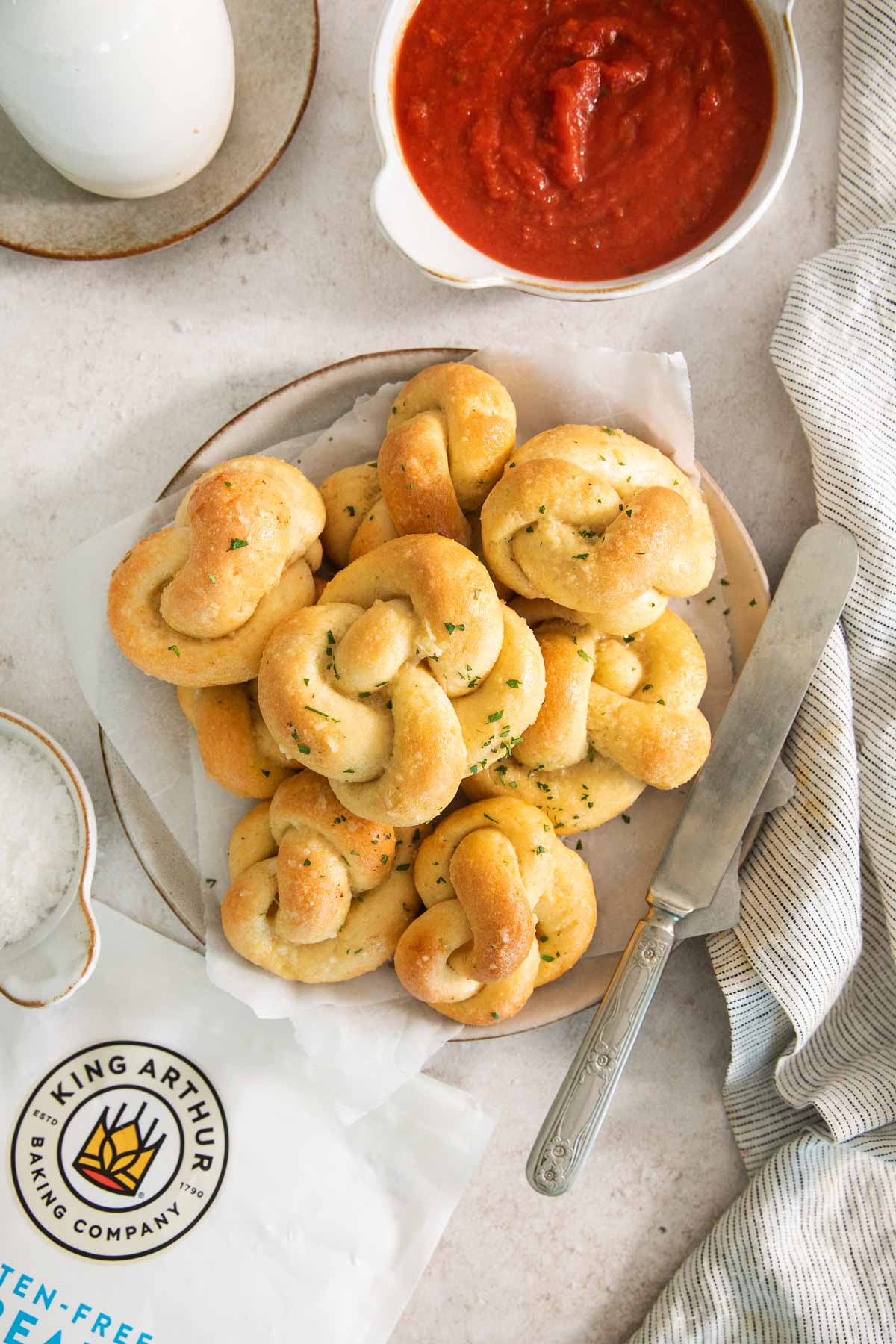 A plate of golden, twisted garlic knots topped with herbs sits on parchment paper next to a bowl of red marinara sauce, a butter knife, a striped napkin, and a King Arthur flour bag.