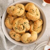 A bowl filled with golden-brown garlic knots sprinkled with chopped parsley, placed on a light-colored table next to a striped cloth.