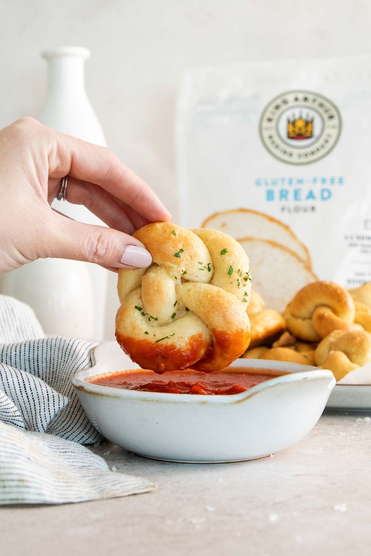 A hand dips a small, twisted soft pretzel into a bowl of marinara sauce. A bag of gluten-free bread flour and several soft pretzels are visible in the background.