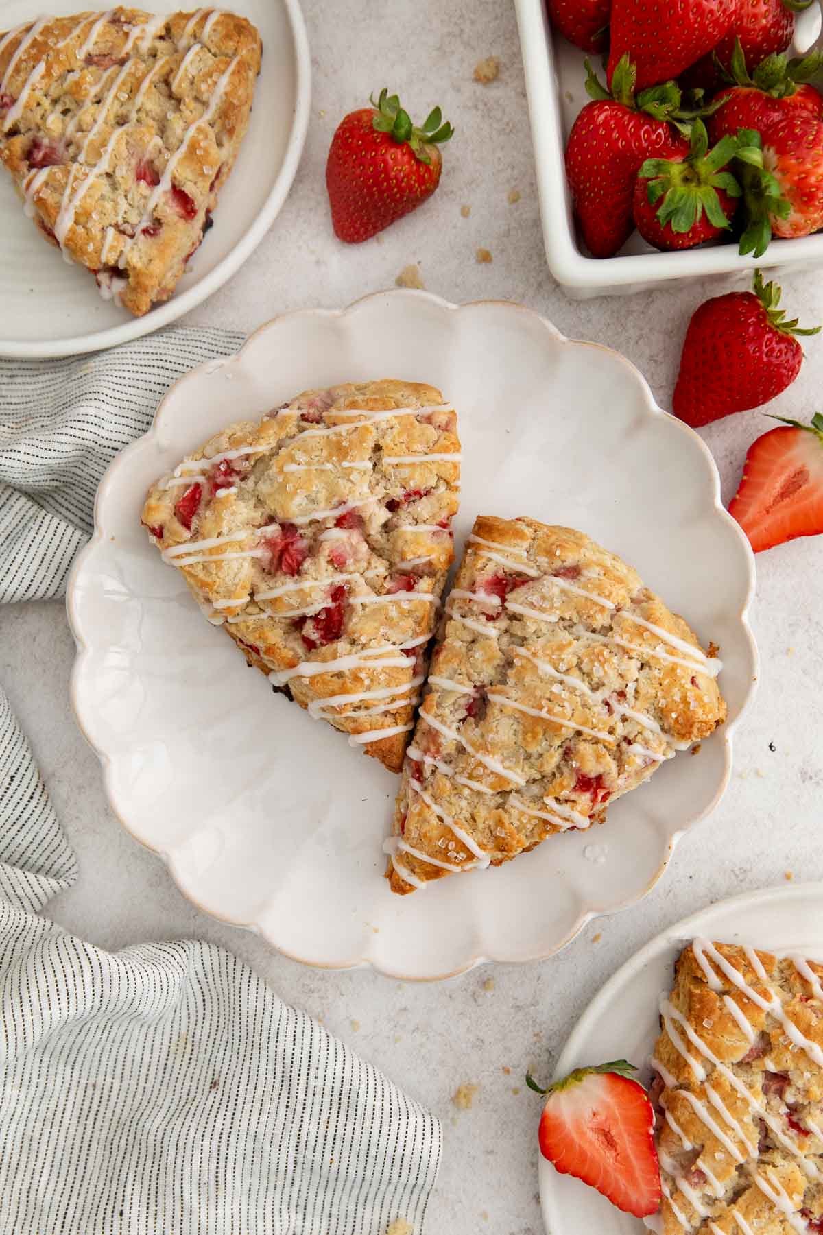 two glazed strawberry scones on a white scalloped plate with more scones and strawberries around.