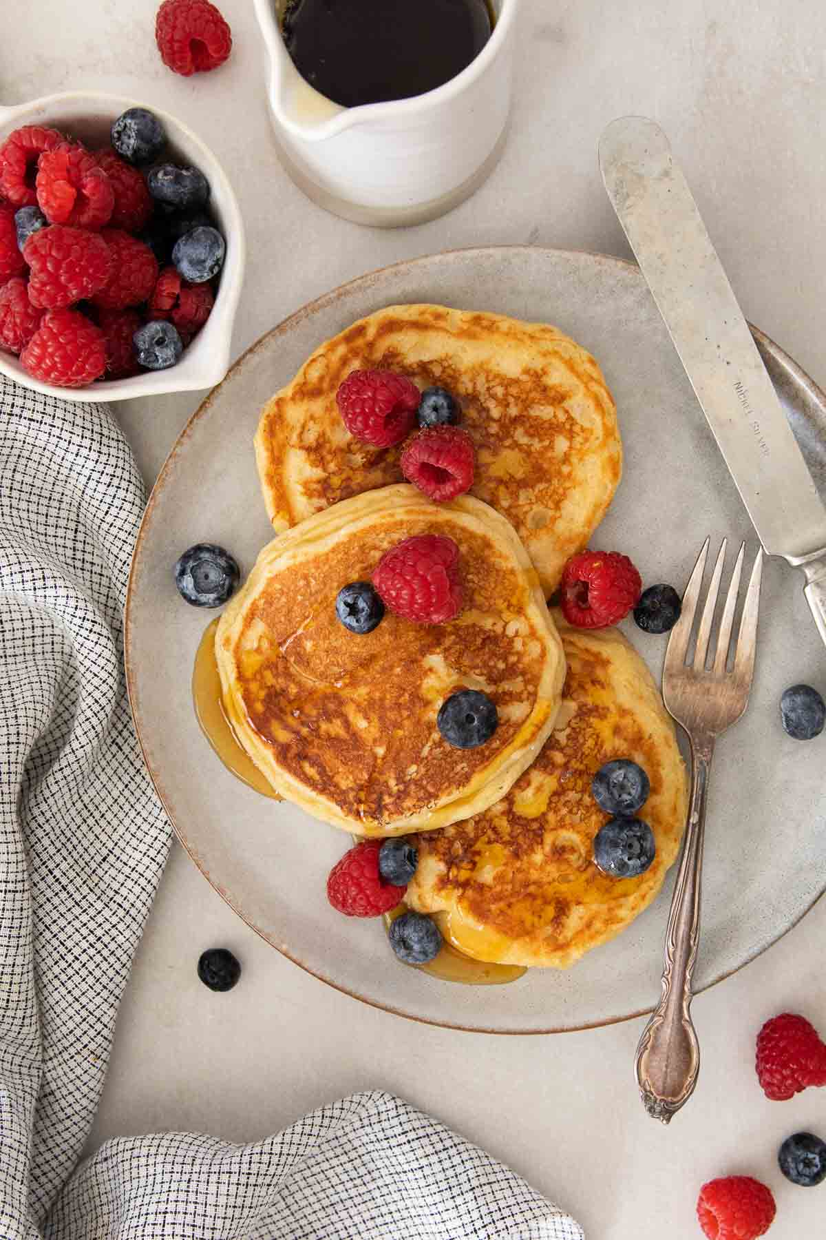 cottage cheese pancakes on a plate with berries, syrup, fork, and a knife.