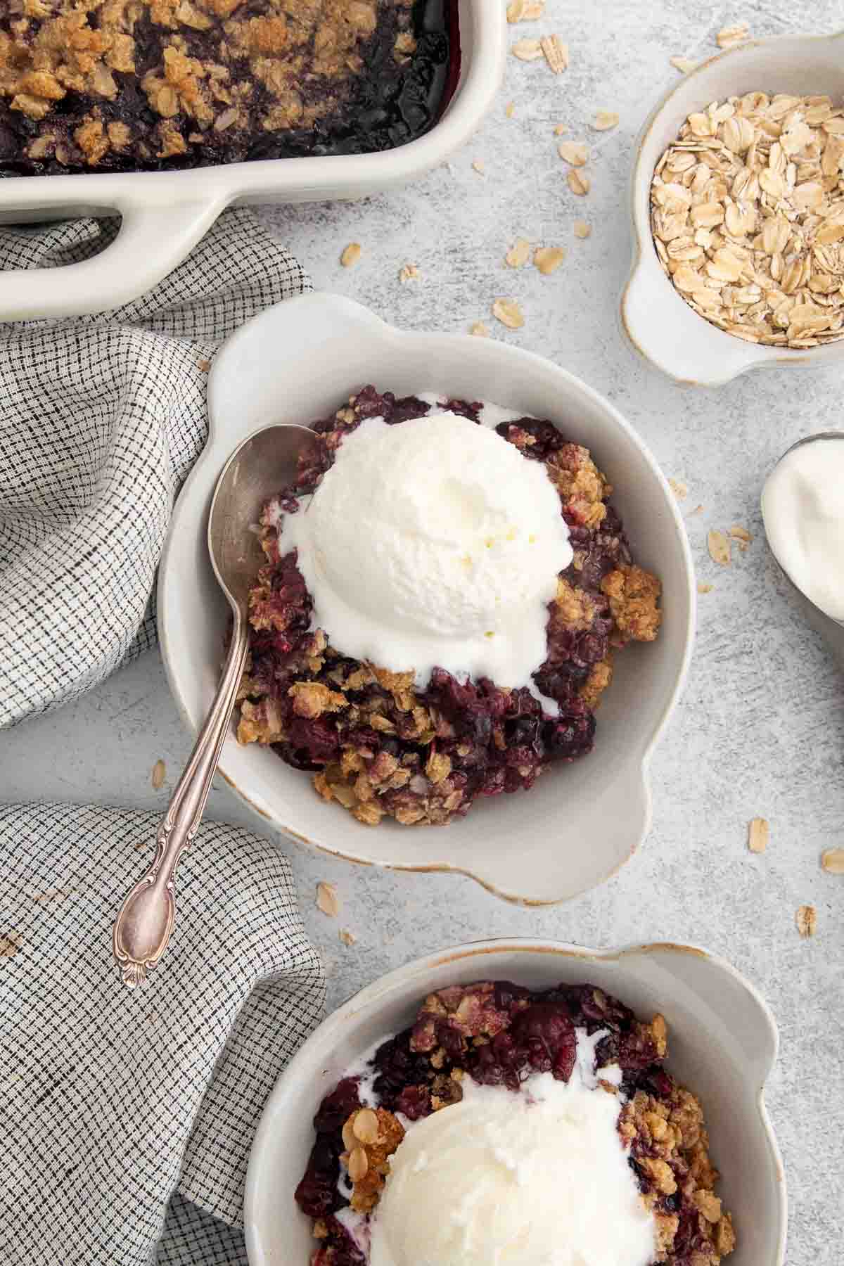 bowls of triple berry crisp topped with ice cream and a spoon next to the dish.