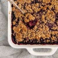 triple berry crisp in a white baking dish with a spoon sticking in it.