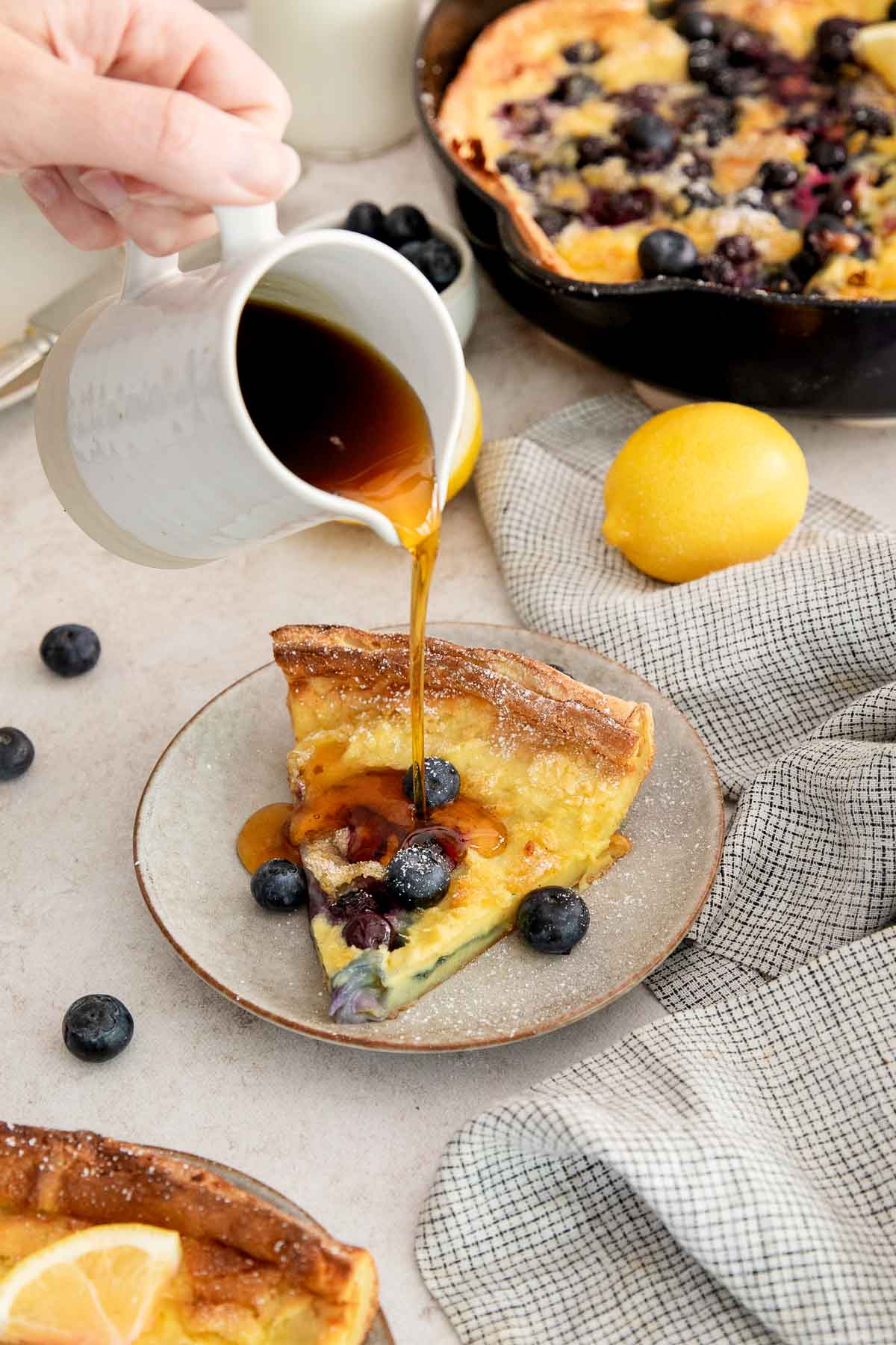 A hand pours syrup over a slice of blueberry Dutch baby pancake on a plate, with fresh blueberries, a lemon, and a skillet of Dutch baby in the background. A gray checked napkin lies nearby.