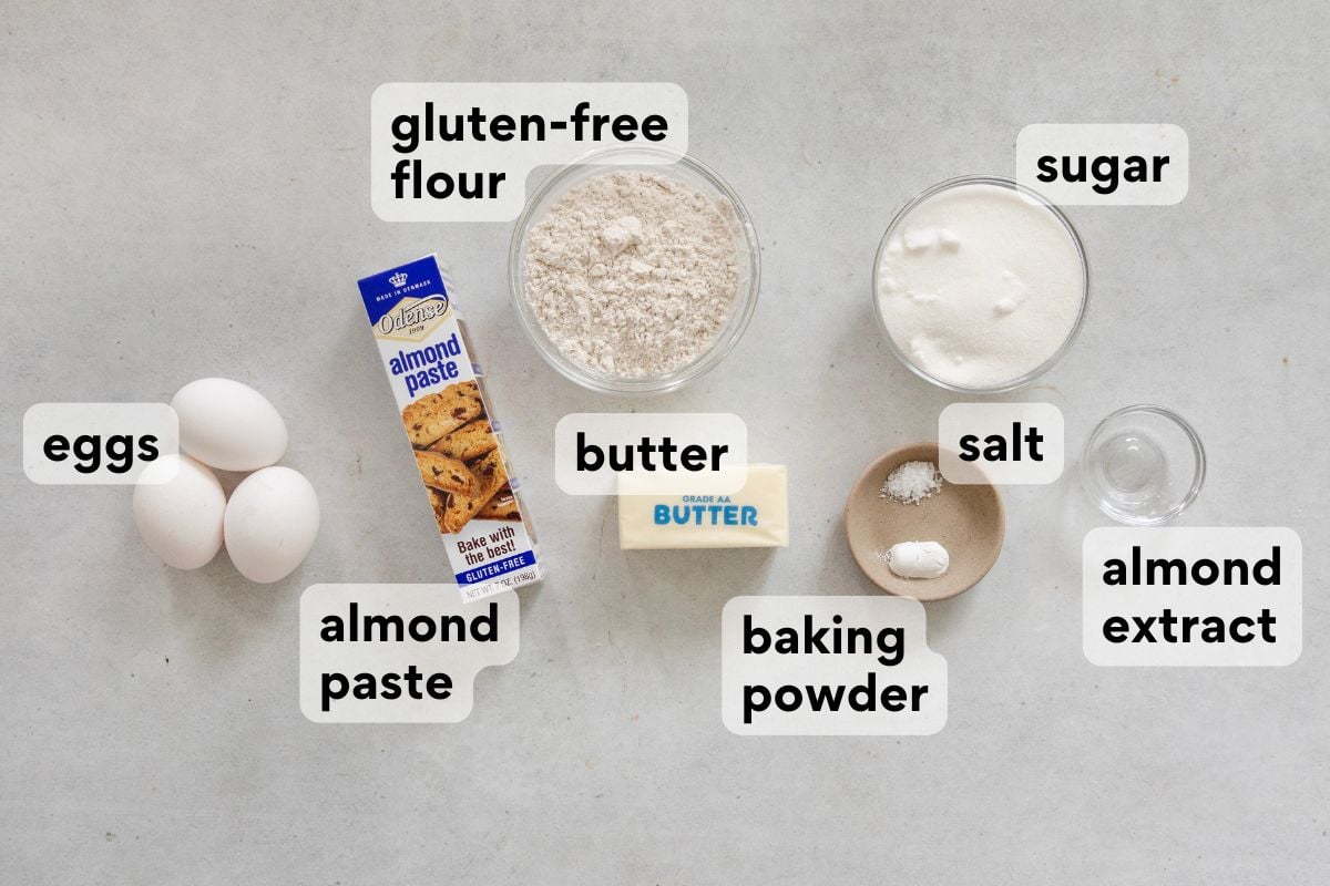 A flat lay of baking ingredients labeled: eggs, almond paste, gluten-free flour, butter, sugar, salt, baking powder, and almond extract, arranged on a light surface.
