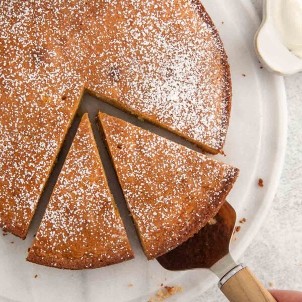 A round, golden-brown cake dusted with powdered sugar sits on a white plate. A slice is being lifted out with a cake server, revealing a moist, tender texture inside.