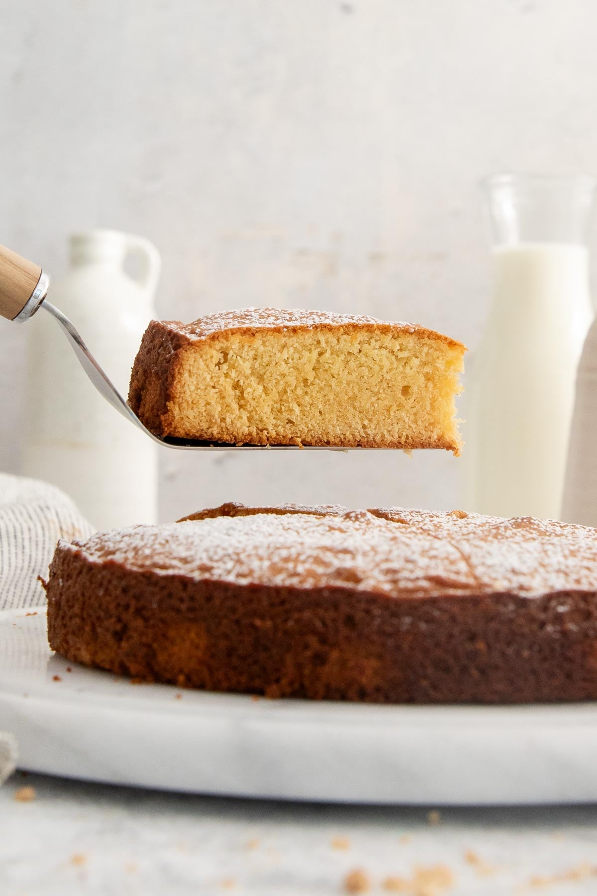 A slice of golden-brown cake is lifted on a spatula above the rest of the cake, which sits on a marble platter. Two bottles of milk are in the background. The cake is lightly dusted with powdered sugar.
