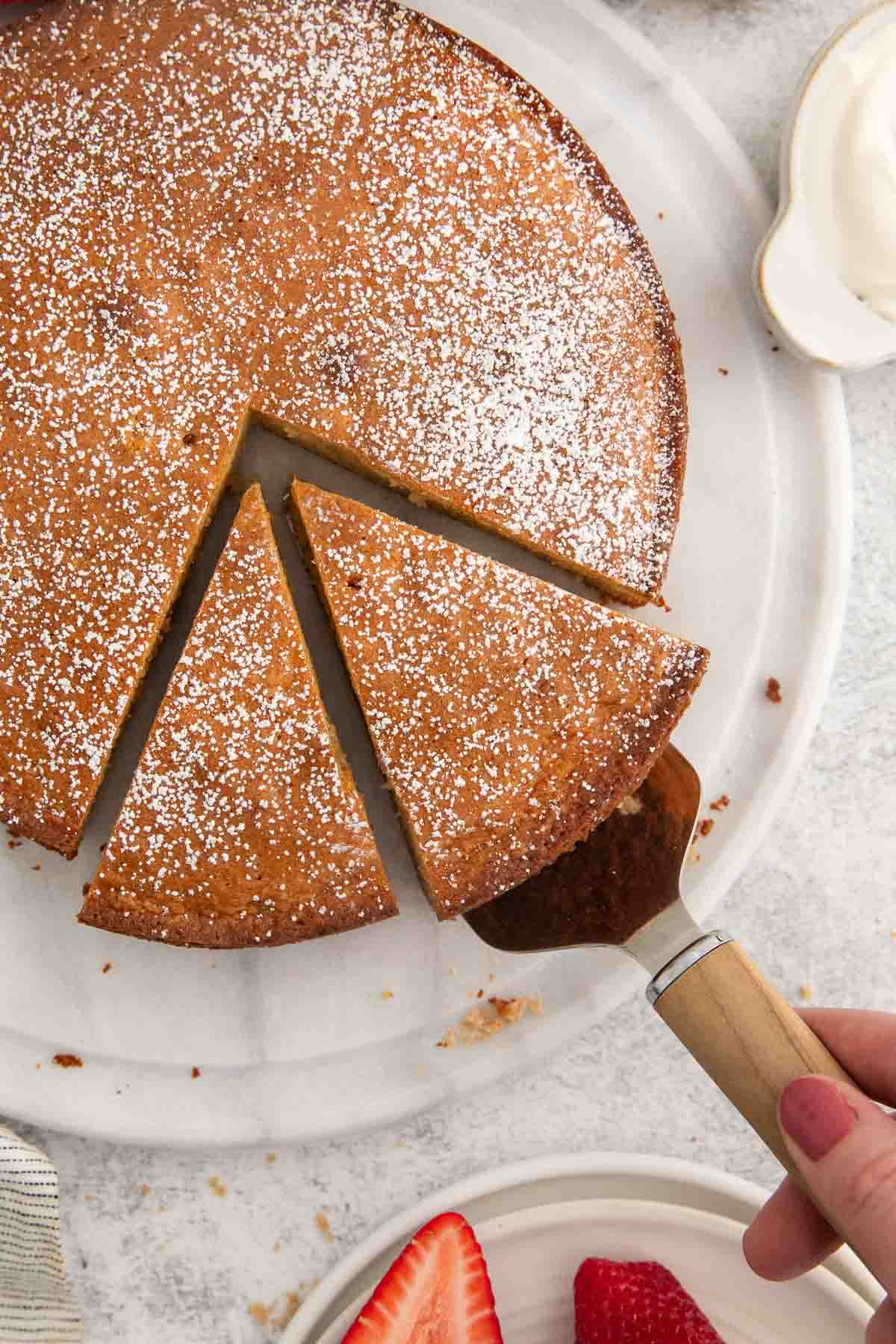 A round cake dusted with powdered sugar sits on a white plate. A slice is being lifted with a wooden-handled server, and fresh strawberries are visible nearby on another plate.