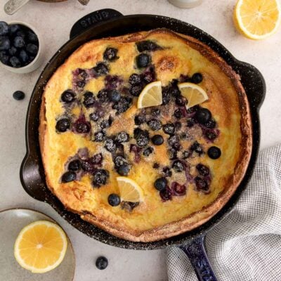 A baked Dutch baby pancake in a cast iron skillet, topped with fresh blueberries, powdered sugar, and lemon wedges. Surrounding the skillet are lemon halves, loose blueberries, a towel, and a bowl of blueberries.