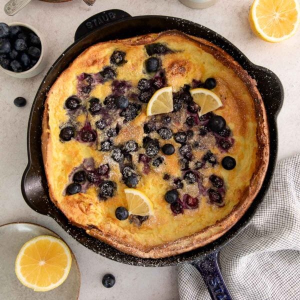 A baked Dutch baby pancake in a cast iron skillet, topped with fresh blueberries, powdered sugar, and lemon wedges. Surrounding the skillet are lemon halves, loose blueberries, a towel, and a bowl of blueberries.