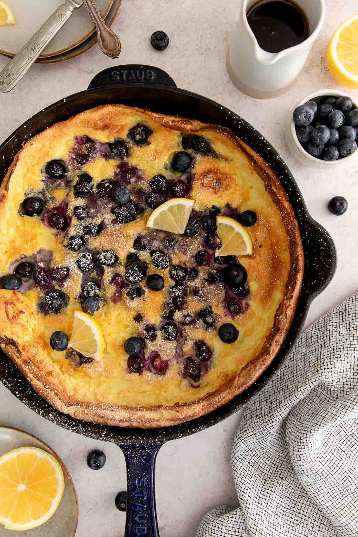 A baked blueberry Dutch baby pancake in a black skillet, topped with fresh blueberries, lemon wedges, and powdered sugar. A small pitcher of syrup, a bowl of blueberries, and a sliced lemon are nearby on a light surface.