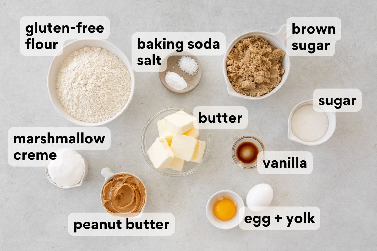 Top-down view of labeled baking ingredients in bowls and plates, including gluten-free flour, baking soda, salt, brown sugar, sugar, butter, vanilla, egg and yolk, peanut butter, and marshmallow creme.